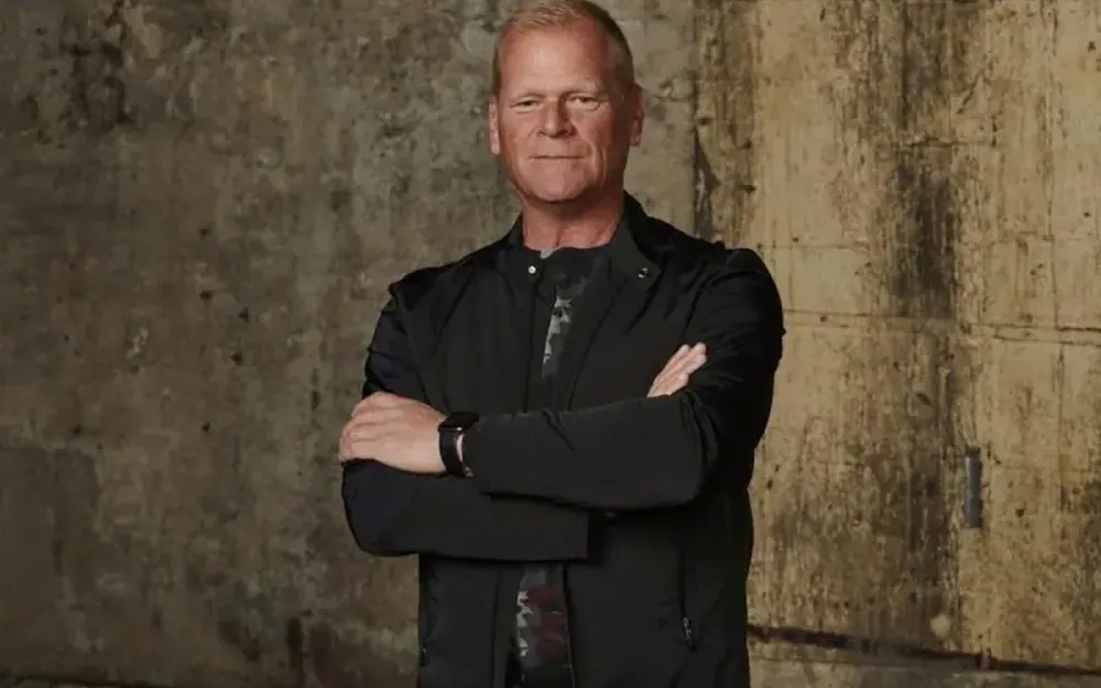 A man in a black jacket standing with arms folded against a concrete wall