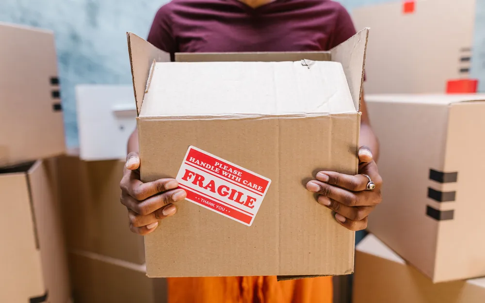 Hands carrying a cardboard moving box marked FRAGILE past a stack of packed boxes